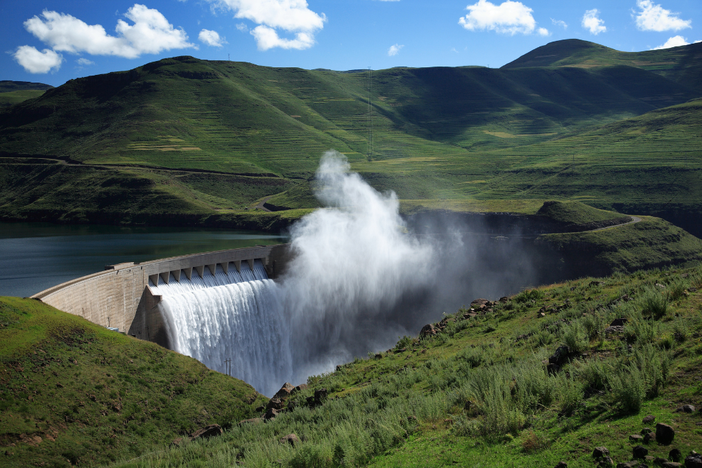 Water gushing from a large dam set in a green mountainous landscape under a blue sky.