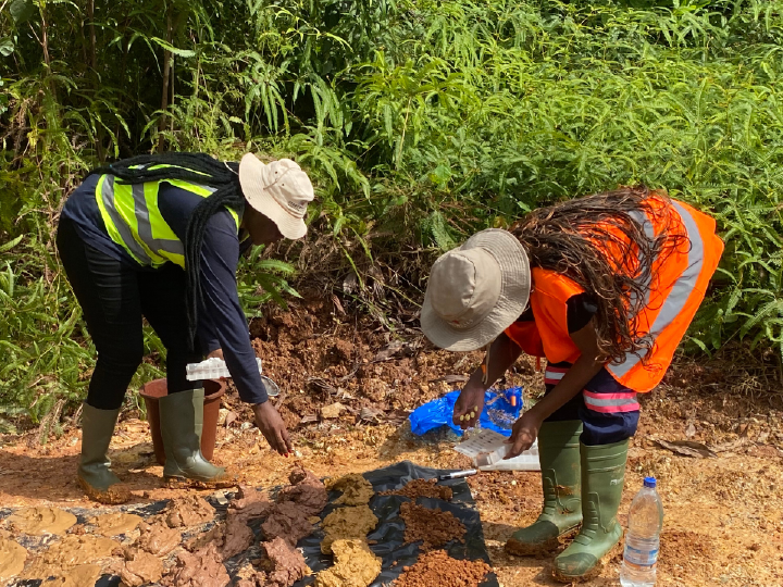 Two female Digby Wells Environmental field scientists — representing the team behind the company's Level 1 B-BBEE status — in high-visibility vests and hard hats, collecting soil samples at an outdoor site surrounded by lush green vegetation.